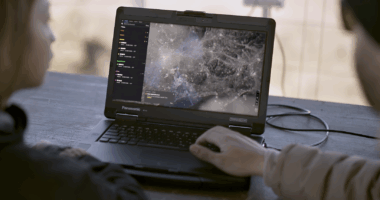 Defense operators using real-time drone surveillance software on a Panasonic Toughbook for mission control and target tracking.