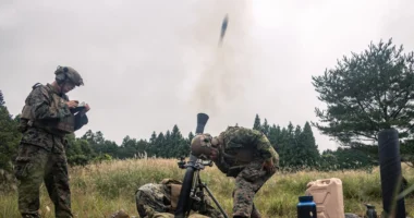 Soldiers conduct a live mortars firing during a field exercise, with one round captured mid-launch from the tube as the crew coordinates targeting and safety procedures in a grassy training area