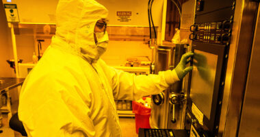 Technician wearing full cleanroom protective gear operates advanced quantum fabrication equipment inside a yellow-lit semiconductor cleanroom, interacting with a control panel on a precision processing system