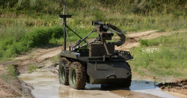 Armed ground robot driving through a muddy dirt road during a field test, equipped with a mounted weapon system and sensors while navigating shallow water in a rural environment