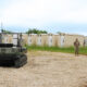Military personnel observe autonomous ground robots operating in a field near temporary structures during a training or testing exercise.