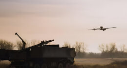 Rheinmetall’s LUNA NG unmanned aerial vehicle flying during a Bundeswehr field trial, operating alongside a ground-based air defense system in a rural training area at dusk
