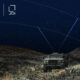 Military vehicle operating at night in rugged terrain, equipped with a satellite terminal linking to overhead satellites for secure communications.