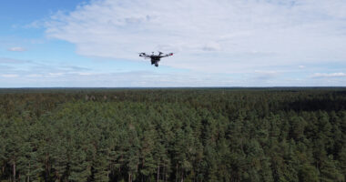 A drone surveys a dense forest under a partly cloudy sky, showcasing how drones can assist in radioactive recovery during daylight hours.