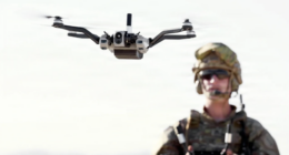 U.S. Army soldier in camouflage and tactical helmet alongside an airborne quadcopter drone equipped with a camera payload, supporting a story on converting vehicle periscope ports into drone launch ports