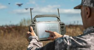 Soldier operating a Getac UX10 rugged ground control station touchscreen in an open field with multiple military drones visible in the background, illustrating a modular unmanned systems command center