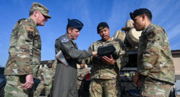 A US Air Force pilot officer and several airmen in OCP uniforms huddle around a handheld tactical device near field equipment, illustrating collaborative efforts to solve emerging drone warfare challenges
