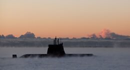 A military submarine emerges through dense sea mist under a dramatic orange dawn sky, used to illustrate Indra's development of a new satellite link to extend submarine connectivity at sea