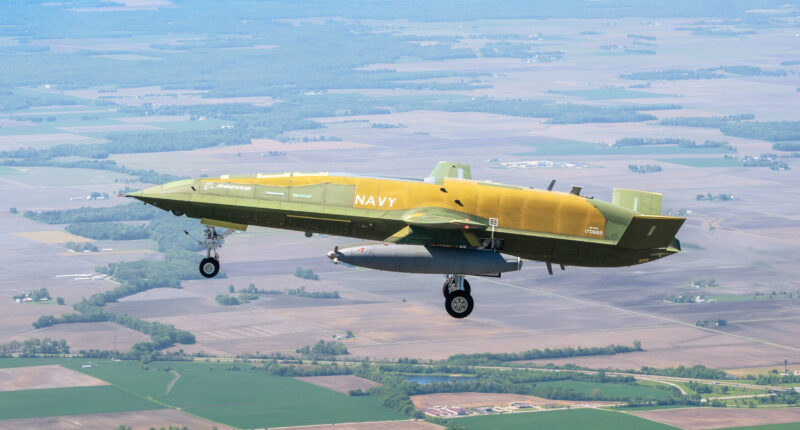 Boeing MQ-25A Stingray unmanned tanker aircraft in green primer and yellow livery during its historic first flight over a rural landscape for the U.S. Navy
