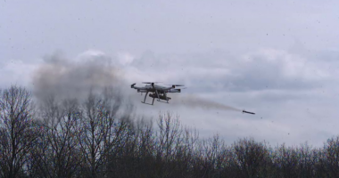 Serbian military autonomous quadcopter drone firing an aircraft rocket with a smoke plume trailing behind it over a bare winter treeline, showcasing the platform's multi-rocket air-to-ground strike capability