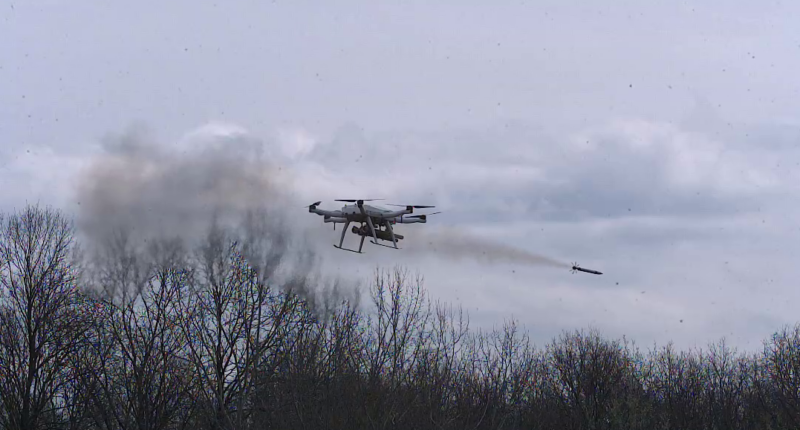 Serbian military autonomous quadcopter drone firing an aircraft rocket with a smoke plume trailing behind it over a bare winter treeline, showcasing the platform's multi-rocket air-to-ground strike capability