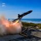 The MGI Defence TigerShark high-speed one-way effector launching from a coastal rail system with rocket booster ignition during test firings.